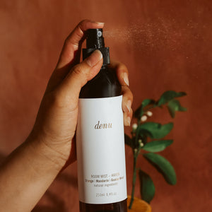 Hand holding a 'denu' Haven room mist bottle with an orange and plant in the background