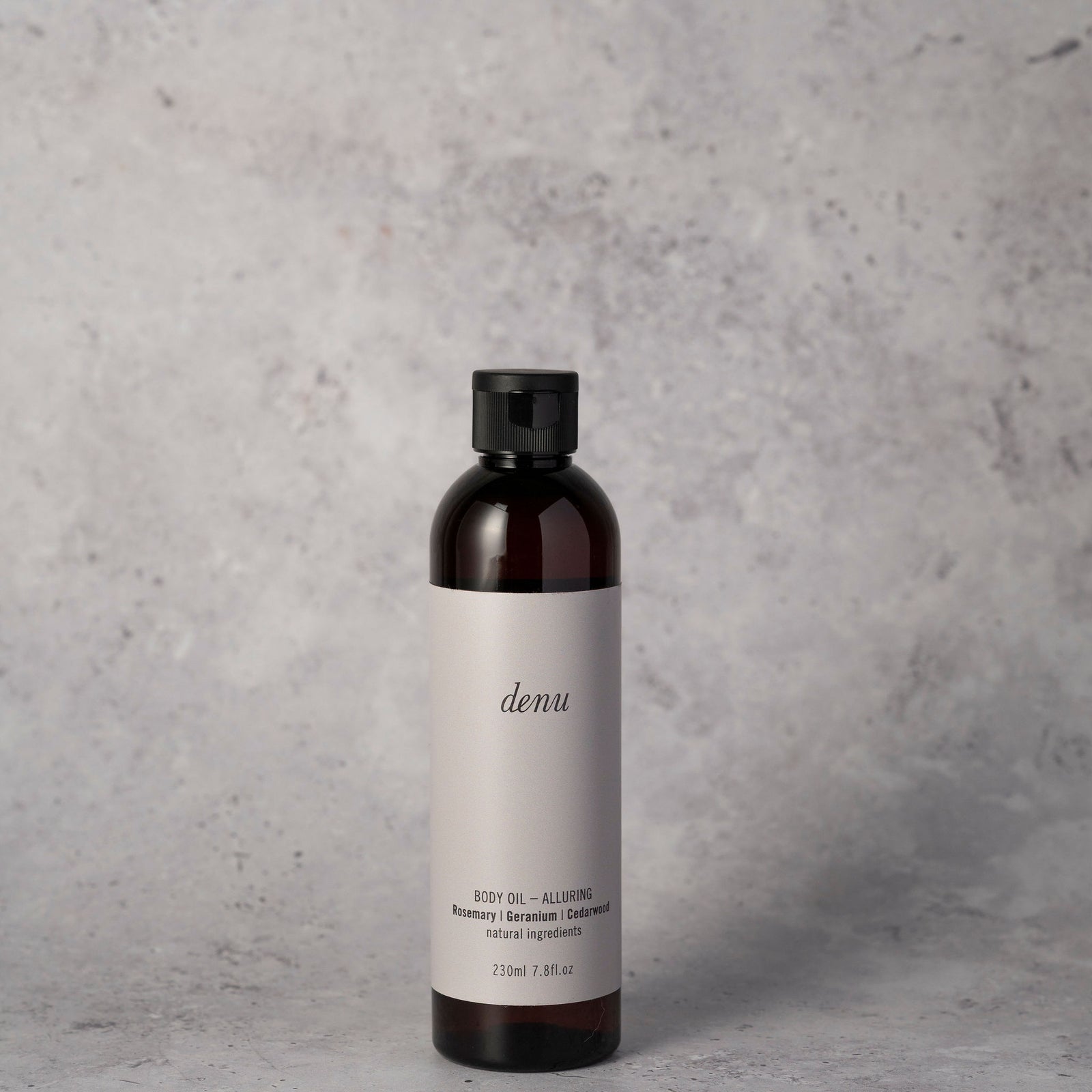 A small dark amber bottle of denu Alluring body oil 50ml with a black cap and a light gray label, on a textured gray surface against a grey background.