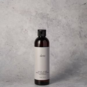 A large dark amber bottle of denu Alluring body oil 230ml with a black cap and a light gray label, on a textured gray surface against a grey background