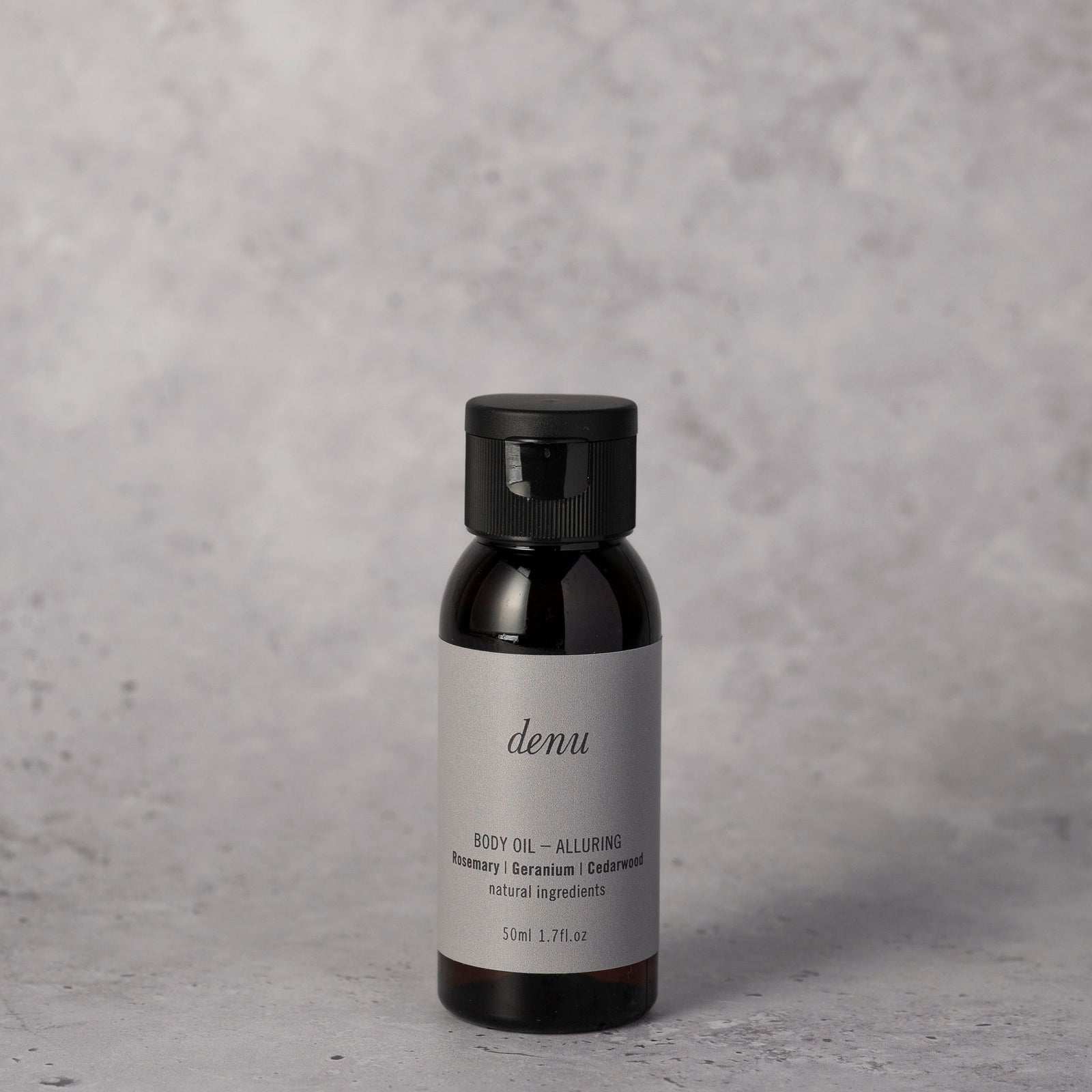 A small dark amber bottle of denu Alluring body oil 50ml with a black cap and a light gray label, on a textured gray surface against a grey background.