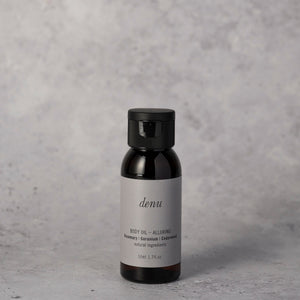 A small dark amber bottle of denu Alluring body oil 50ml with a black cap and a light gray label, on a textured gray surface against a grey background.